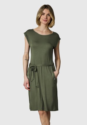 Frau trägt ein knielanges olivgrünes Kleid mit kurzen Ärmeln, einem Taillengürtel und seitlichen Taschen, steht vor einem hellgrauen Hintergrund.