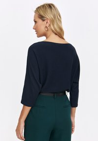 Granatowy sweter z rękawami o długości trzech ćwiartek, luźny fason i szeroki dekolt, w połączeniu z zielonymi spodniami o wysokim stanie z tylnymi kieszeniami.