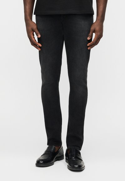Homme portant un jean noir slim et des chaussures à enfiler en cuir noir, debout devant un fond clair uni.