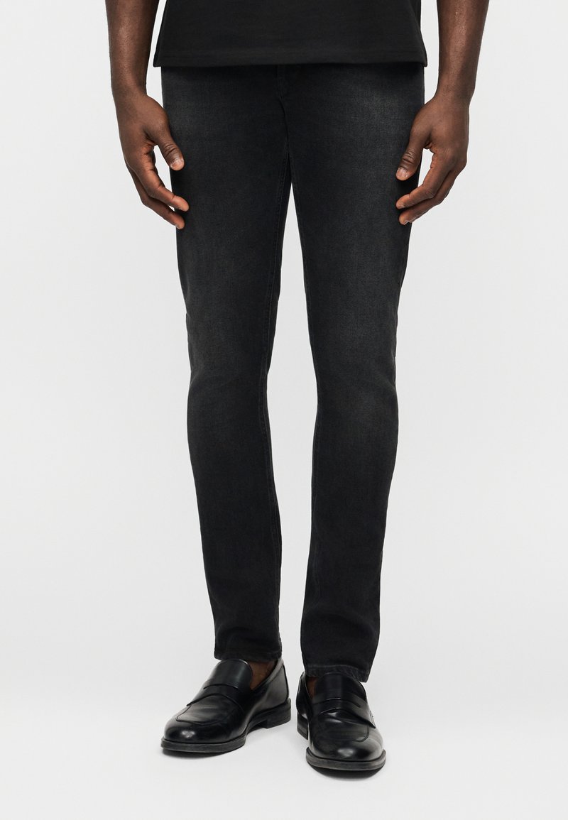 Homme portant un jean noir slim et des chaussures à enfiler en cuir noir, debout devant un fond clair uni.