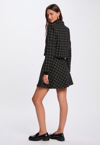 Giacca corta nera a quadri con collo alto, abbinata a una gonna nera coordinata. Il tessuto ha una finitura strutturata e dettagli metallici.