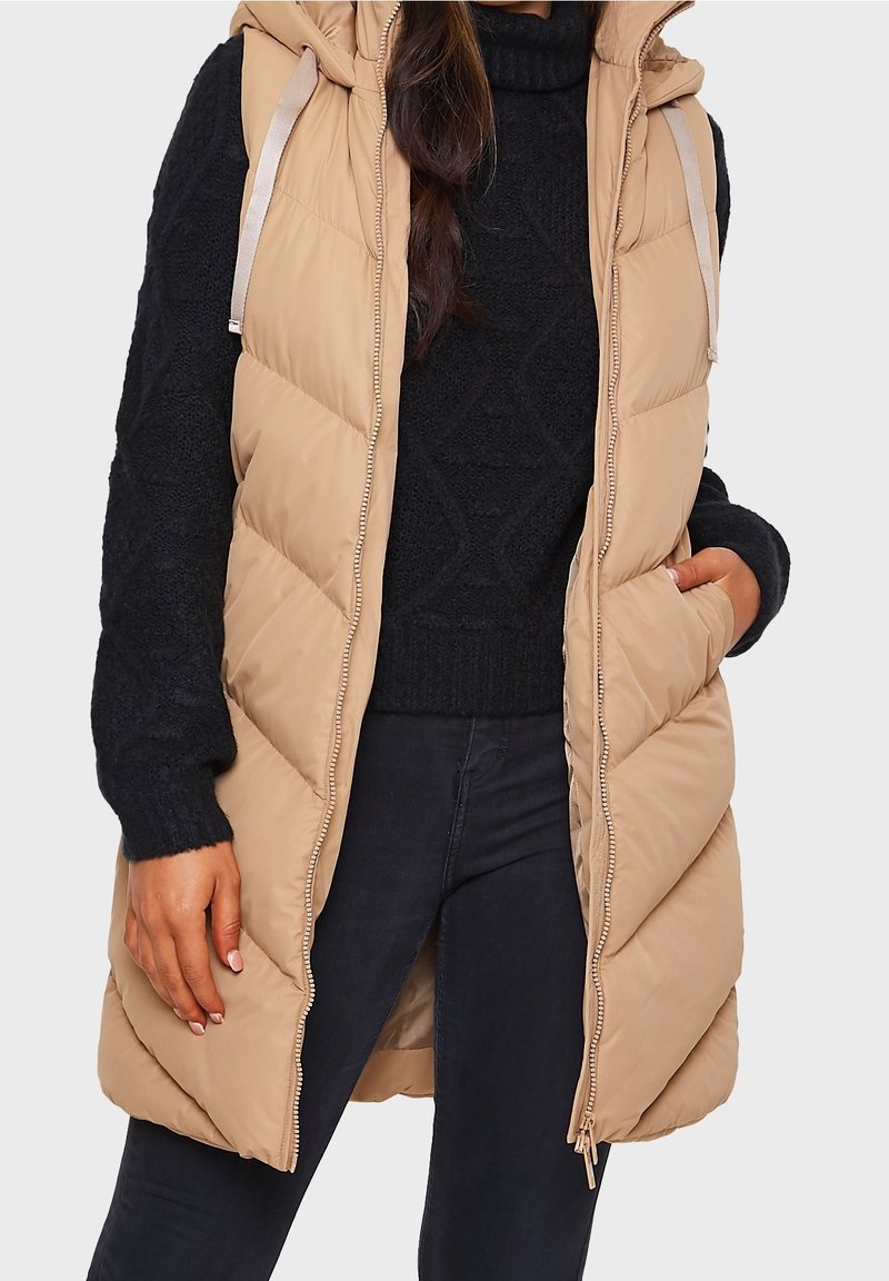 Beige gepolsterte Weste mit Kapuze, diagonal gestepptem Muster, Reißverschluss vorne und silbernen Hardware-Akzenten. Über einem dunklen Pullover getragen.