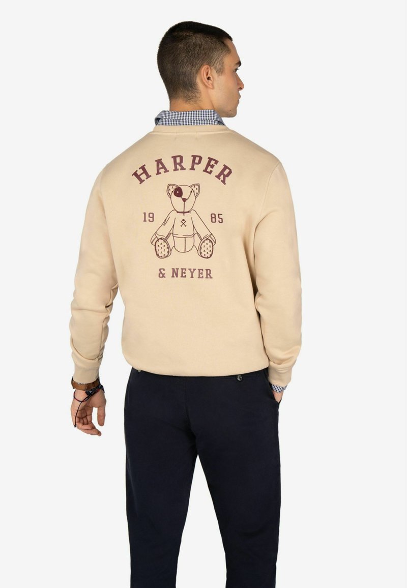 Sudadera beige con un gráfico de un oso de dibujos animados, "HARPER" y "1985" impresos en burdeos. Presenta puños y cuello acanalados.