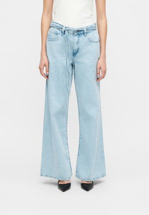 Person trägt hellblaue Jeans mit weitem Bein und angenähtem Jeansgürtel sowie schwarze spitze Schuhe, steht vor einfarbigem Hintergrund.