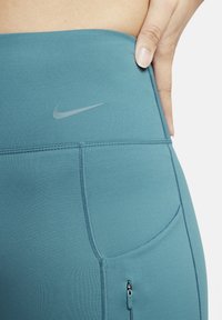 Legginsy Nike z wysokim stanem w tealowym kolorze charakteryzują się gładką fakturą, srebrnym logo oraz akcentowanym zamkiem błyskawicznym w kieszeni z boku.