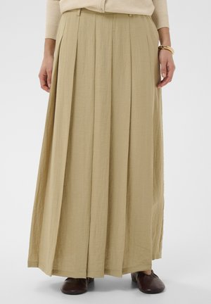 Beżowa plisowana spódnica maxi noszona z beżową bluzką, brązowymi butami i bransoletkami na prawej ręce, widoczna od talii do stóp.