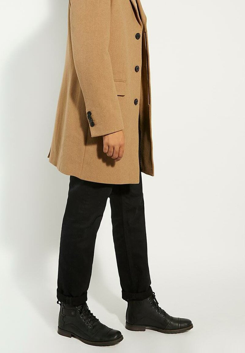 Camel wool blend coat with black buttons, paired with black tapered pants and black lace-up boots. The coat has side pockets and a structured fit.