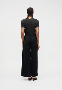 Άτομο με πλεξούδες, φορώντας εφαρμοστή μαύρη μπλούζα, μαύρο παντελόνι φαρδιάς γραμμής και μαύρα παπούτσια με τακούνι, στέκεται με την πλάτη προς το μέρος μας σε λευκό φόντο.