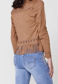 Veste en daim marron avec franges et détails ajourés floraux portée avec un jean bleu clair brodé, vue de dos et de côté.