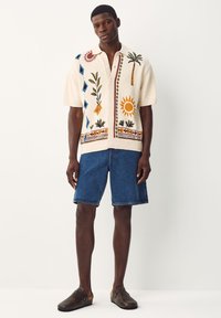 Kortærmet, cremefarvet striktrøje med farverig broderi, herunder sol, palmer og geometriske mønstre, kombineret med denimshorts og brune træsko.