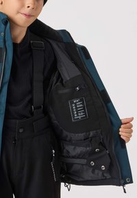 Wodoodporna kurtka w kolorze morskim z czarnym wnętrzem. Posiada liczne kieszenie, regulowane paski oraz metkę z instrukcjami pielęgnacji i informacjami o funkcjach.