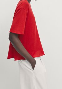 T-shirt rouge à manches courtes en tissu doux, présentant une coupe ample et un ourlet arrondi, associé à un pantalon blanc.