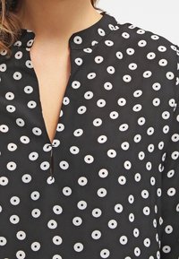 Blusa nera con scollo a V e motivi circolari bianchi. Realizzata in un tessuto liscio con una lavorazione morbida.