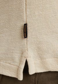 Beige strukturierter Pullover mit einem kurzen Seitenschlitz, versehen mit einem kleinen schwarzen Etikett mit der Aufschrift "PME Legend." Das Material wirkt weich und gewebt.