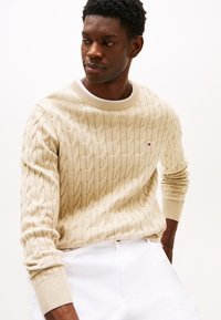 Μπεζ πουλόβερ με πλέξη καλώδιου, λαιμόκοψη τύπου crew και ριμπ μανσέτες. Διαθέτει διακριτικές λεπτομέρειες μοτίβου και μικρό λογότυπο στο στήθος.
