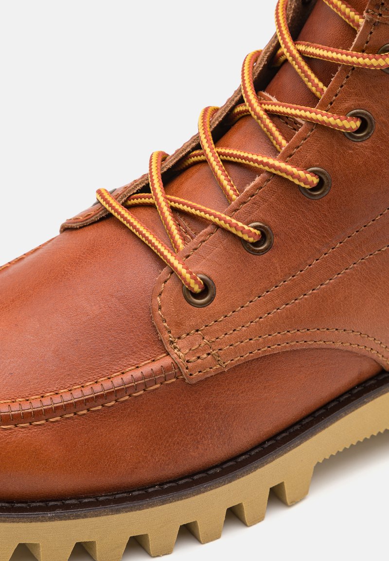 Acercamiento de una bota de cuero marrón con cordones amarillos y rojos en un patrón y suela de goma beige gruesa con profundas acanales.