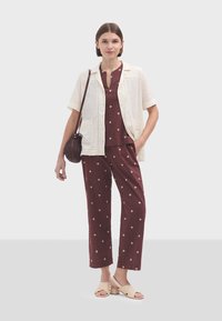 Chemise légère et boutonnée sur un haut bordeaux, avec un pantalon à motifs. Sandales à talon épais. Sac rond et texturé.