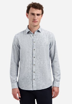 Mann trägt ein hellgraues, strukturiertes Hemd mit langen Ärmeln und Knöpfen sowie dunkle Hosen vor weißem Hintergrund.