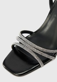 Melnas papēža sandales ar kvadrātu formas purngalu, gludu ādas tekstūru un vairākiem joslu zandaliem, kas rotāti ar caurspīdīgiem rhinestones.