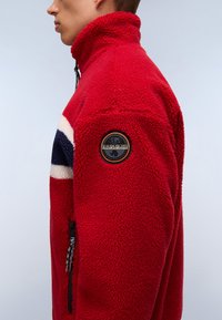 Jachetă din fleece roșie cu guler înalt, având dungi bleumarin și crem. Include o buzunare lateral cu fermoar și un patch rotund cu logo pe mânecă.
