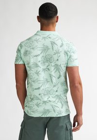 Vaaleanvihreä poolopaita, jossa on tummanvihreä kukka- ja lintukuosi. Lyhyet hihat ja klassinen kaulus, valmistettu hengittävästä kankaasta.