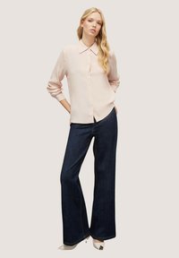 Camicia button-up rosa chiaro con colletto, abbinata a pantaloni a gamba larga blu scuro. L'outfit presenta una texture liscia e colori in contrasto.
