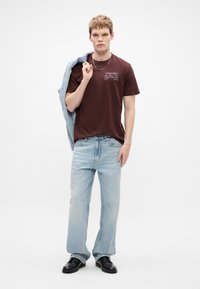 Camiseta de algodón marrón con texto impreso en blanco, jeans azul claro y zapatos de cuero negros. Una chaqueta de mezclilla clara está colgada sobre un hombro.