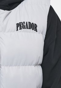 Vit pufferväst med svart "PEGADOR" broderi på bröstet, med en slät textur och kviltad design, lagd över en mörk topp.
