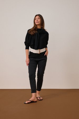 Vrouw staat blootsvoets, draagt een zwarte trui, zwarte broek, brede witte riem, met de trui over de schouders gedrapeerd tegen een effen achtergrond.