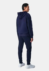 Felpa navy con cappuccio e pantaloni jogger in tessuto morbido; presenta un cappuccio, polsini a coste e un logo sulla manica. Abbinato a sneaker bianche.
