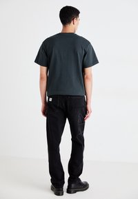 Dark green cotton t-shirt with short sleeves, paired with black denim pants featuring multiple pockets and a label tag. Black footwear completes the look.