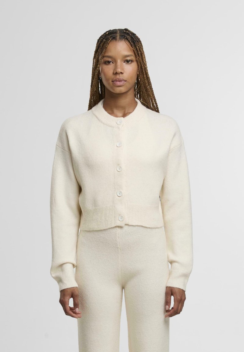 Kremowy krótki kardigan z okrągłym dekoltem, z przodu zapinany na guziki, z prążkowanymi mankietami i dołem, wykonany z miękkiej, fakturowanej tkaniny.