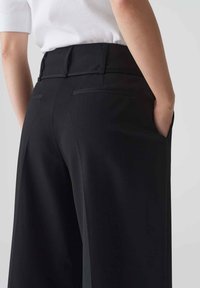 Zwarte wijde broek met een gladde textuur, een getailleerd ontwerp en verborgen achterzakken. Aanpassingen aan de tailleband zijn zichtbaar.
