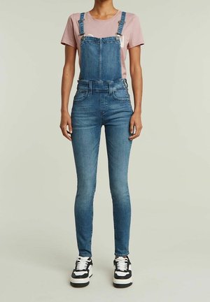 Denim overalls in a medium blue wash, with adjustable straps, a bib pocket, and slim tapered legs. Paired with a light pink t-shirt and black sneakers.
