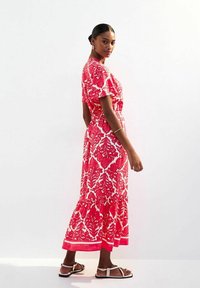 Maxikleid mit rot-weißen Blumenmuster, kurzen Ärmeln, Taillenknoten und gerafftem Saum. Getragen mit flachen Sandalen.