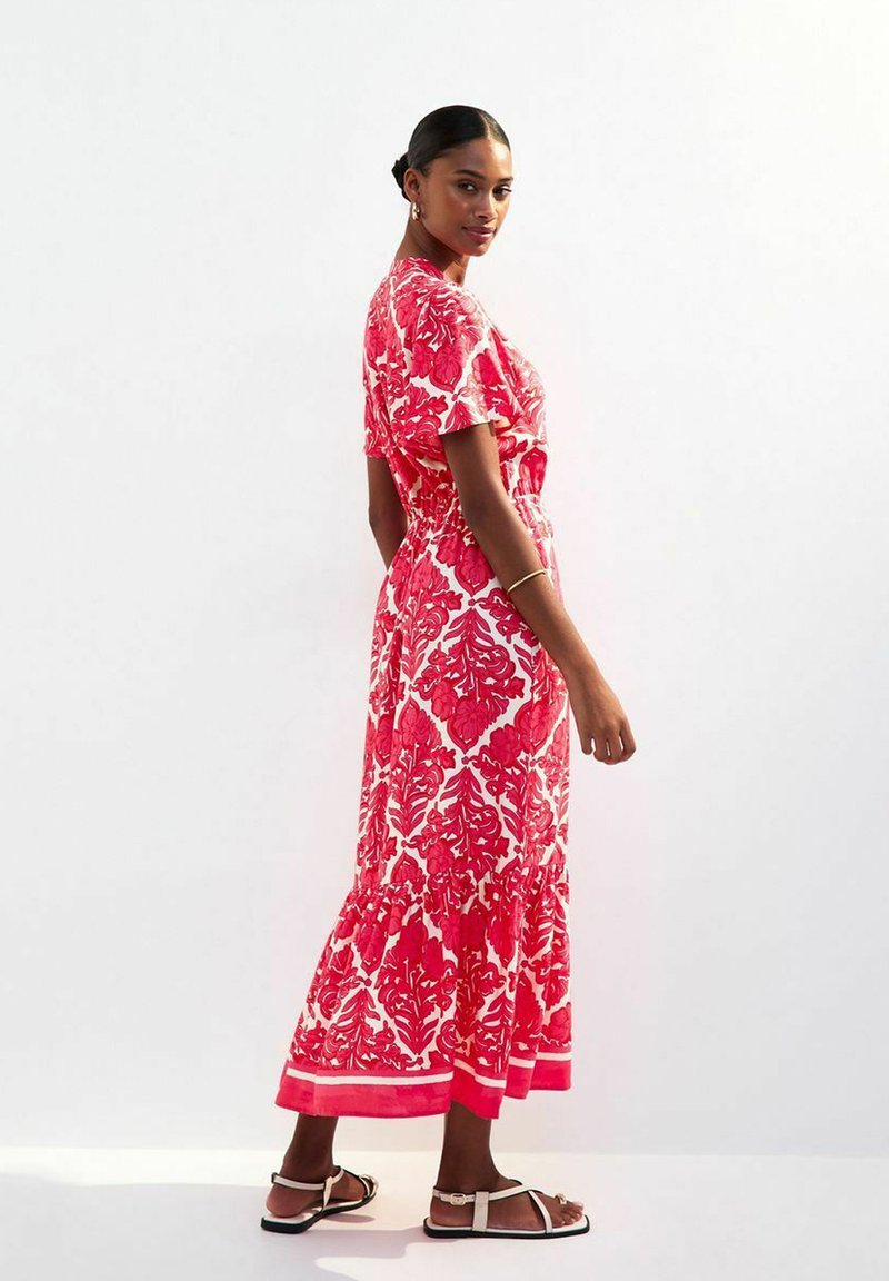 Robe maxi à motifs floraux rouges et blancs avec des manches courtes, une ceinture à la taille et un ourlet volanté. Portée avec des sandales plates.