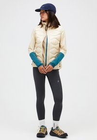 Gilet imbottito crema con dettagli turchesi, maglia a maniche lunghe, leggings scuri e scarpe da trekking beige. Un cappellino casual completa l'outfit.