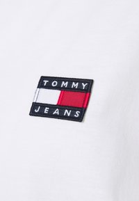 Vitt bomullstyg med en sydd rektangulär logotyp. Logotypen har blå, röd och vit section med texten "TOMMY JEANS" i svart.