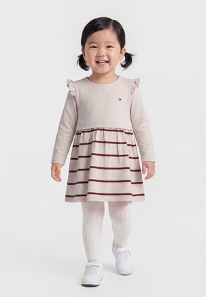 Junges Mädchen mit Zöpfen, das ein beiges Kleid mit roten Streifen, weiße gemusterte Strumpfhosen und weiße Turnschuhe trägt, lächelnd und nach vorne gehend.