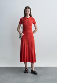 Mujer que lleva un vestido midi rojo de manga corta con botones y patrones texturizados, con un pequeño bolso de hombro plateado y zapatos Mary Jane negros.