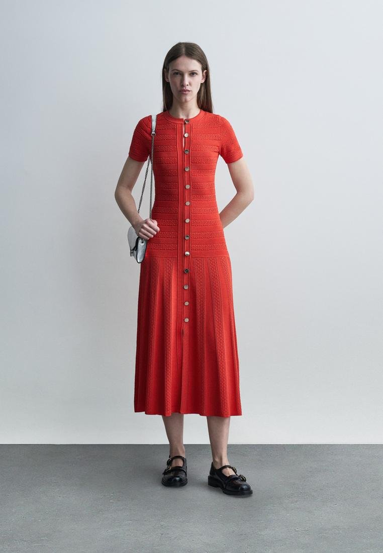 Femme portant une robe midi rouge à manches courtes boutonnée avec des motifs texturés, portant un petit sac à bandoulière argenté et des chaussures Mary Jane noires.
