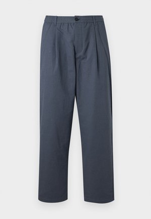 Homme à la tête rasée portant un t-shirt blanc, une chemise rayée claire ouverte, un pantalon foncé avec ceinture et des sandales noires, debout les mains dans les poches.