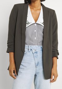 Olivgrön blazer med uppvikta ärmar över en vit skjorta med vågig krage och svart rutig mönster, kombinerad med ljusblå högmidjade jeans.