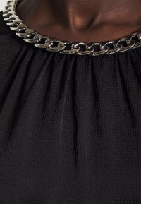 Gros plan sur un collier à chaîne argentée porté sur une peau foncée, au-dessus d'un vêtement en tissu noir texturé avec des détails froncés.