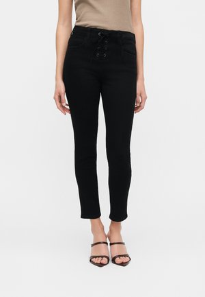 Femme portant un jean skinny noir taille haute avec un détail à lacets sur le devant et des talons ouverts noirs, debout devant un fond uni.