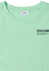 T-shirt vert clair avec col rond, étiquette "CECIL" et texte noir disant "SELFCARE Wellness n'est pas une tendance. C'est un investissement en soi."