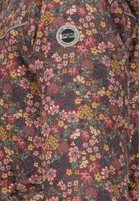 Ärmel einer Jacke mit dichtem, mehrfarbigem Blumenmuster auf burgunderfarbenem Stoff, versehen mit einem runden "Zigzag" Markenpatch.