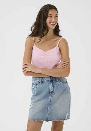 Mladá žena sa usmieva so založenými rukami, oblečená v svetloružovej camisole a svetlomodrej rifľovej sukni s ošúchaným okrajom.