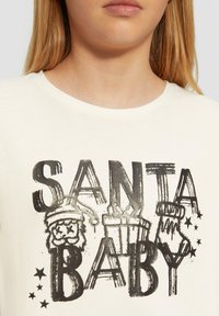 Witte katoenen t-shirt met een zwarte grafische print waarop "SANTA BABY" staat, vergezeld van illustraties van een kerstmanhoed, cadeaus en sterren.