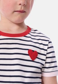 Ριγέ μπλουζάκι με κόκκινο λαιμόκοψη και κοντά μανίκια. Διαθέτει υφασμάτινη κόκκινη καρδιά στο αριστερό στήθος πάνω σε λευκές και ναυτικές ρίγες.